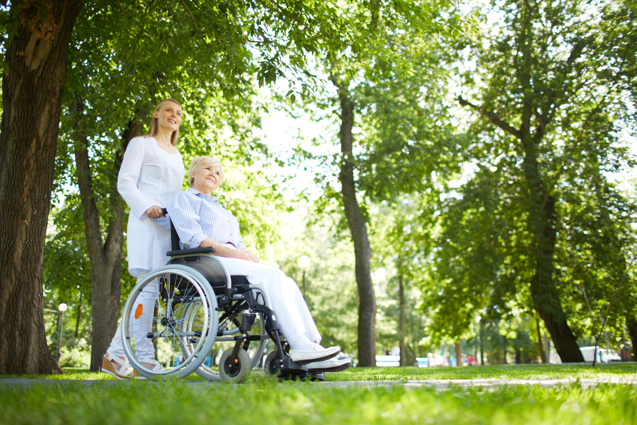 A woman providing Clayton senior home care services pushes an older woman in a wheelchair through the park.