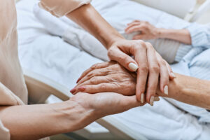 A woman providing care for her mother who has Alzheimer’s at the end of life gently holds her mother’s hand as she rests in bed.