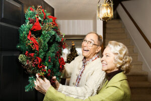 A senior couple hang a holiday wreath on their front door because decorating is a good way to create holiday joy for loved ones with dementia.